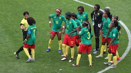 Mehrere Fußball-Nationalspielerinnen von Kamerun stehen zusammen mit einer Schiedsrichterin im Mittelkreises des Platzes beim WM-Spiel gegen England. © imago images 