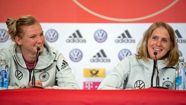DFB-Nationalspielerin Alexandra Popp (l.) und Co-Trainerin Britta Carlson lächeln während einer Pressekonferenz auf dem Podium. © dpa picture alliance Foto: Sebastian Gollnow