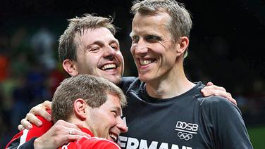 Die deutschen Tischtennisspieler Timo Boll (Mitte), Bastian Steger (l.) und Trainer Jörg Rosskopf (r.) jubeln. © dpa Foto: How Hwee Young